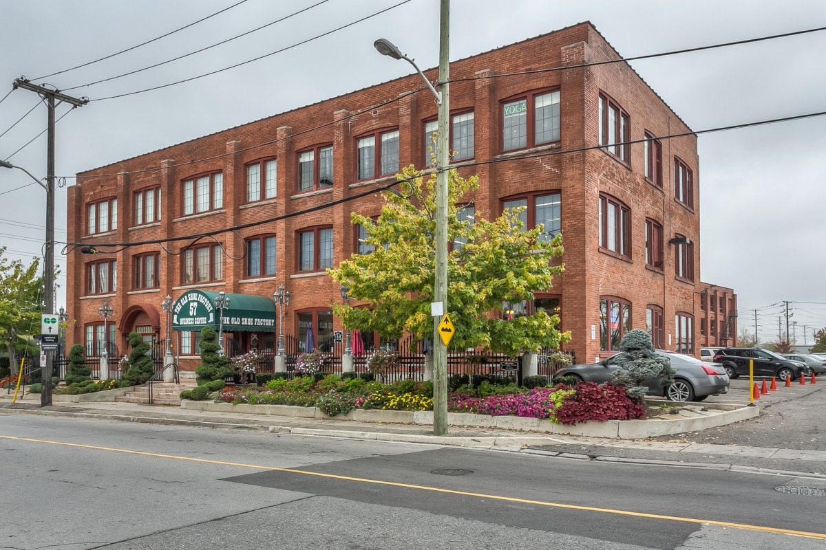 Historic red brick building, The Old Shoe Factory, features lush landscaping and a yoga studio sign, showcasing a blend of heritage and modern wellness.