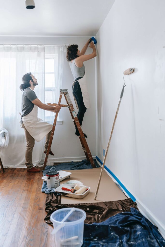 A woman stands on a ladder painting the wall while a man supports her. Tools and paint materials are spread on the floor, indicating a home renovation project.