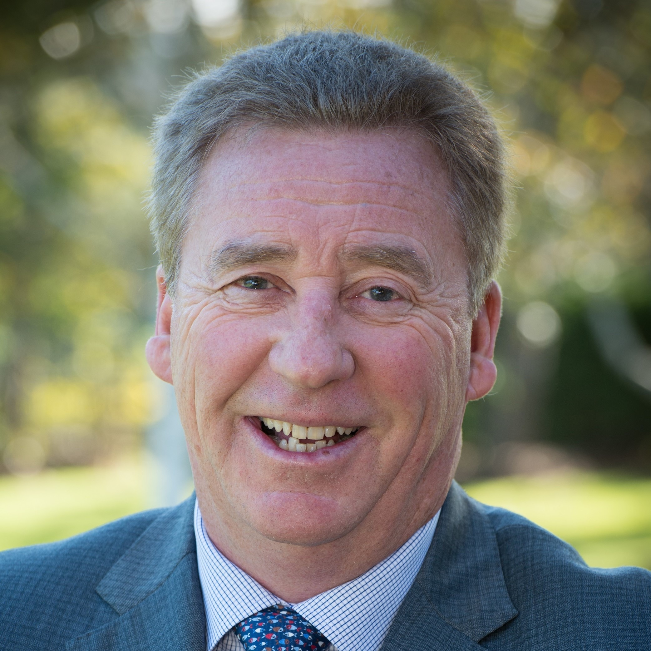 Smiling man in a suit outdoors, with blurred greenery in the background, conveying a friendly and approachable demeanor.