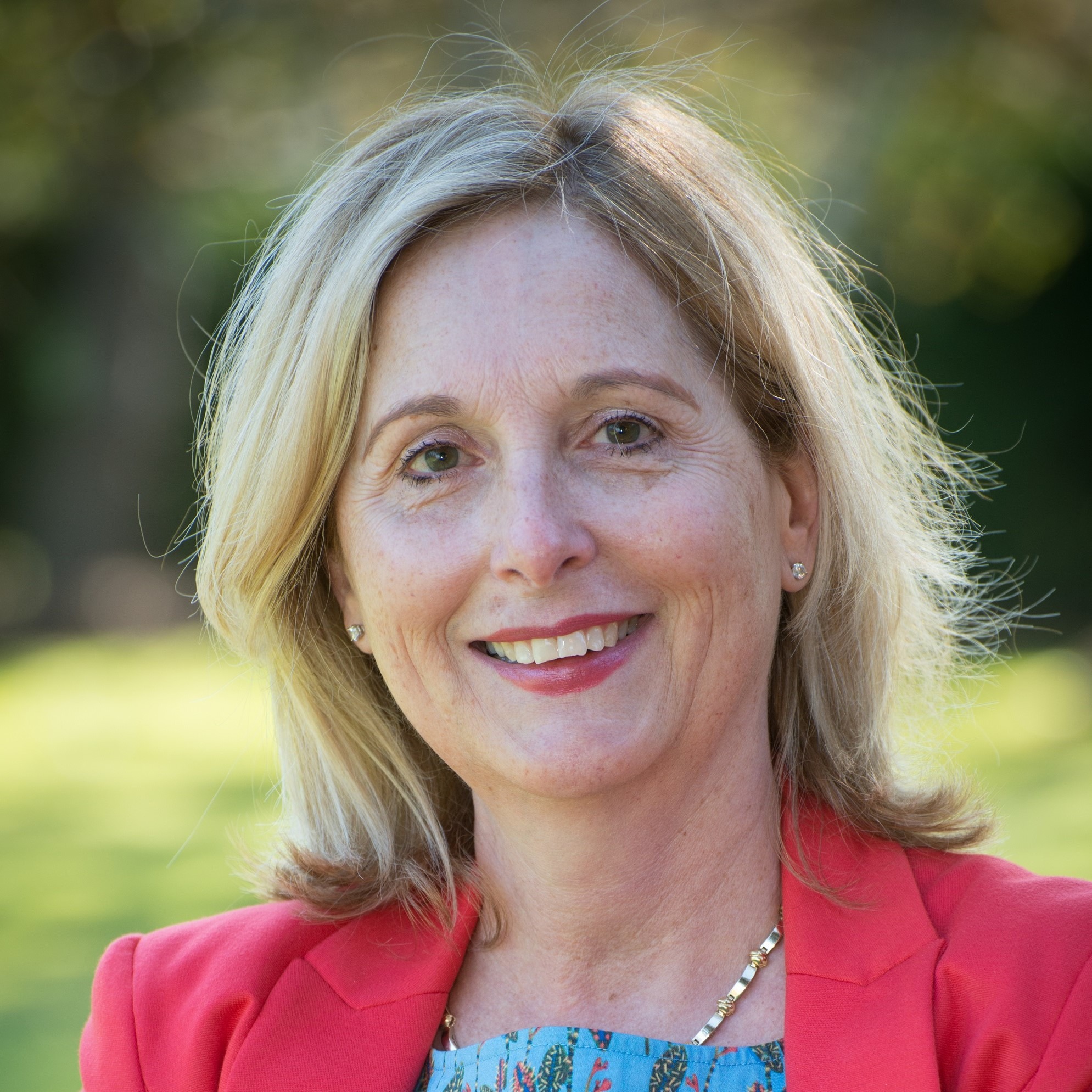 Smiling woman with blonde hair in a red blazer and floral blouse, set against a blurred green background, exuding warmth and confidence.