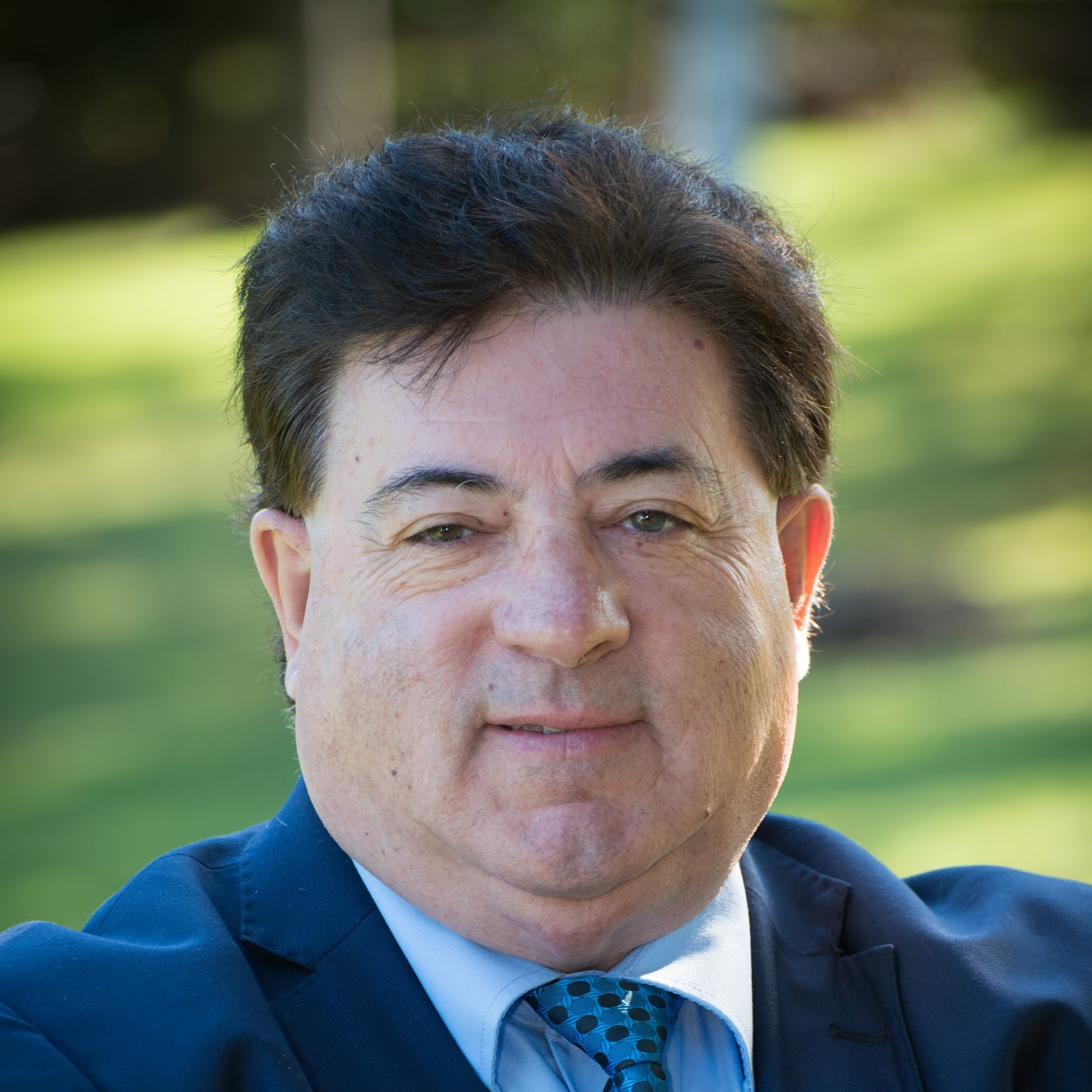 A man in a suit and tie smiles confidently against a blurred green background, conveying professionalism and approachability.