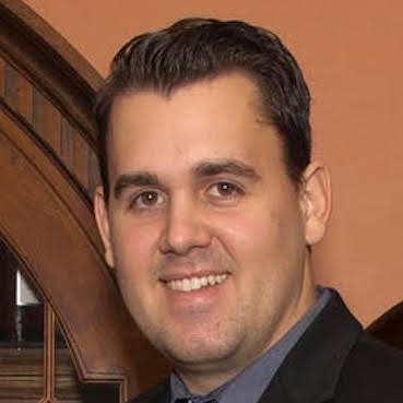 A smiling man in a formal black suit poses in front of an ornate wooden backdrop, representing professionalism and approachability.