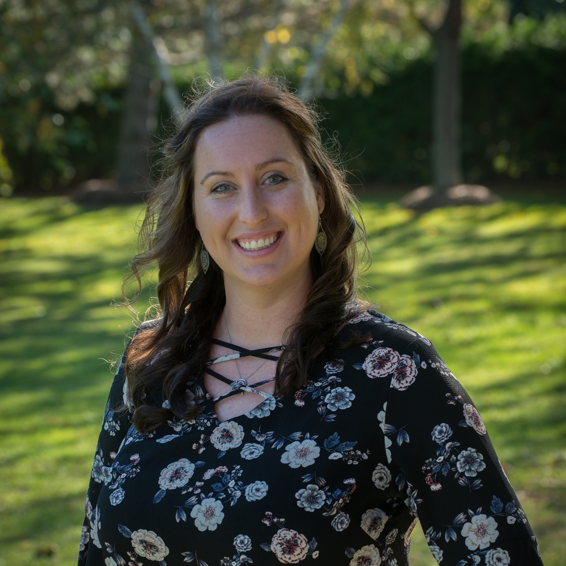 A smiling woman with long brown hair stands outdoors in a floral-patterned top, against a backdrop of green grass and trees, exuding warmth and approachability.