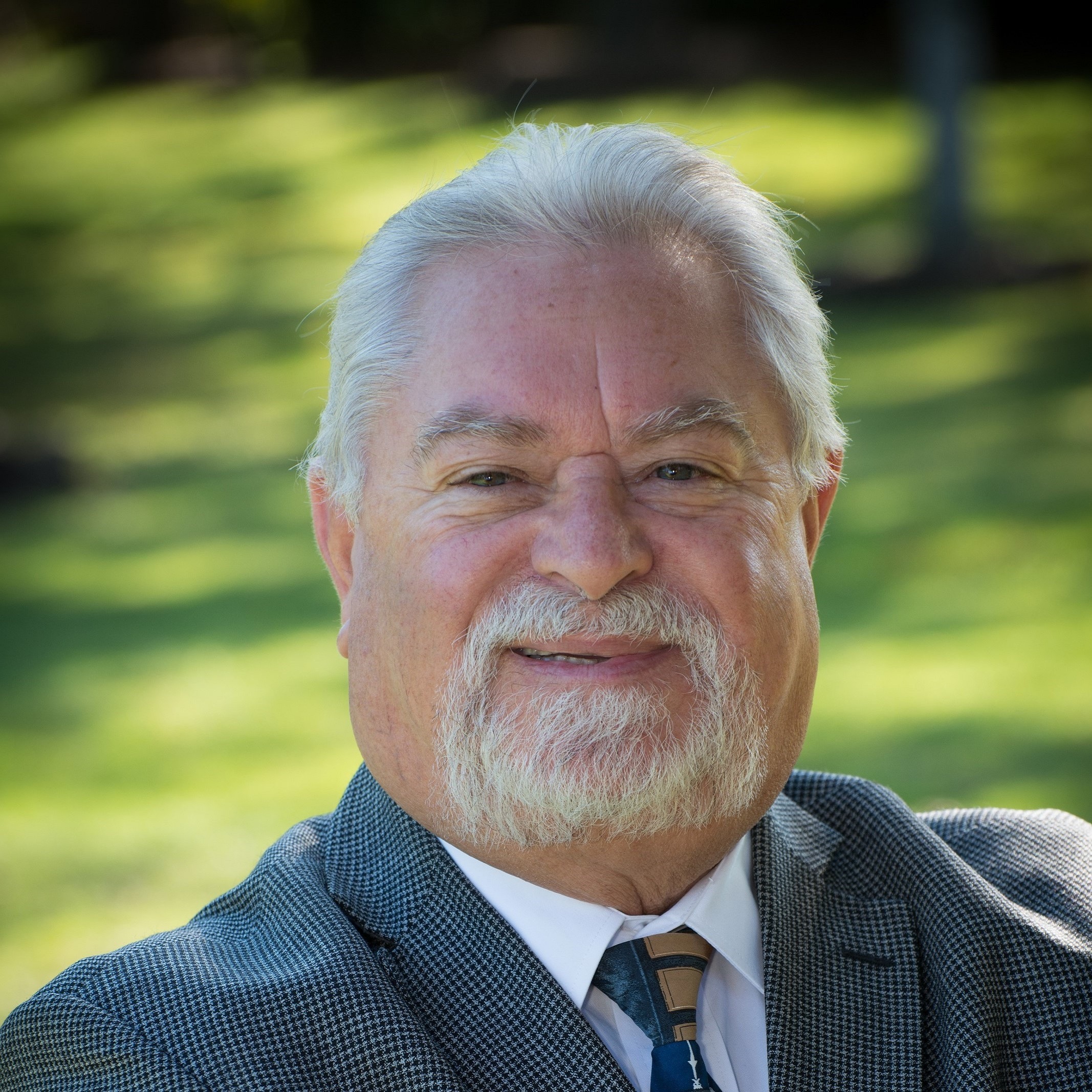 Smiling older man in a suit with a patterned tie, posed outdoors against a green blurred background. Suitable for professional networking or biography.