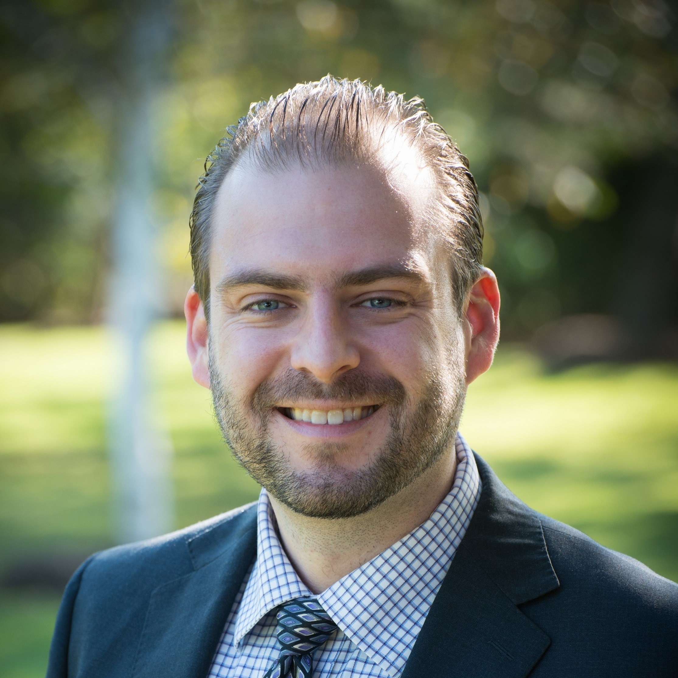 Smiling man in a suit and tie stands outdoors, with blurred greenery in the background. His confident expression suggests a professional context.