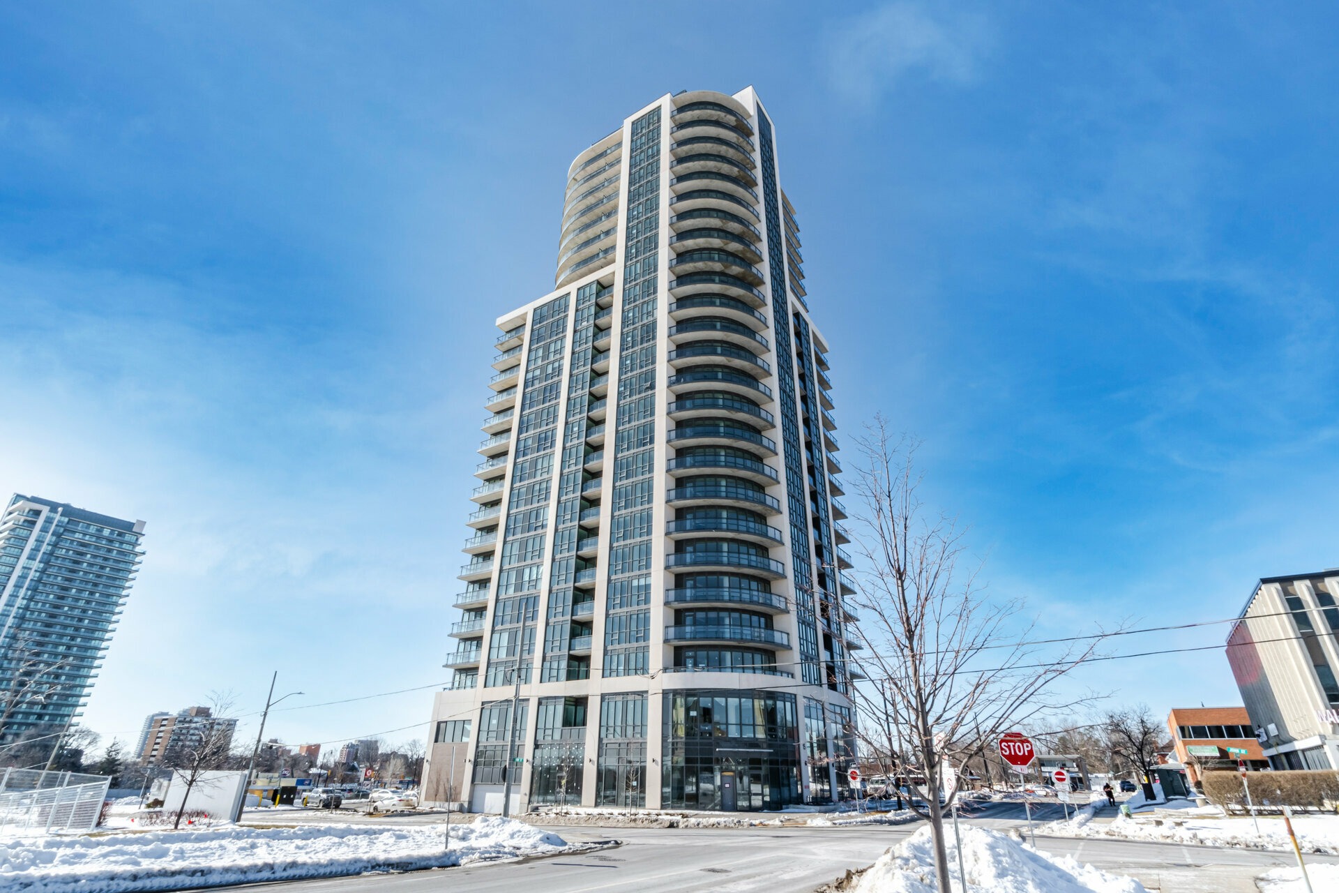 Modern high-rise condominium tower showcases sleek glass balconies against a clear blue sky. Snow blankets the surrounding area, highlighting urban development.