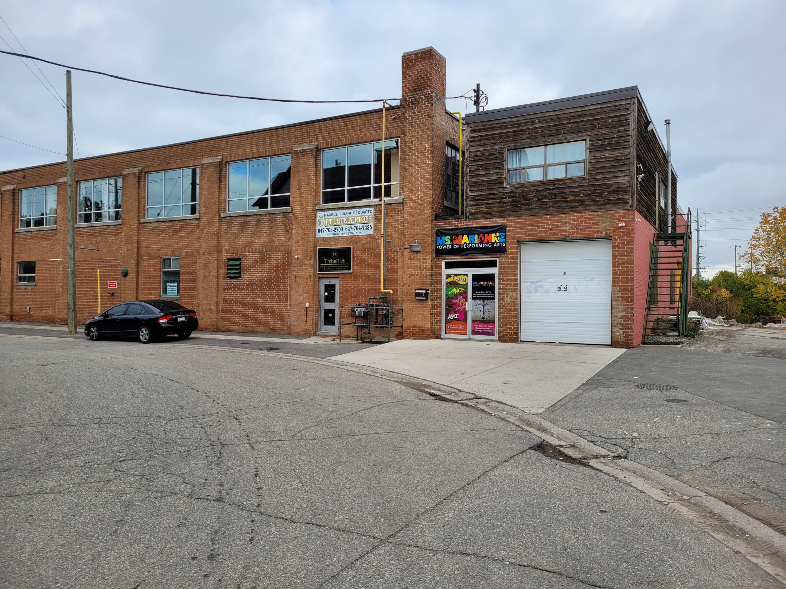 Brick building with large windows and a signage for a performing arts center. A black car is parked nearby on a paved street.