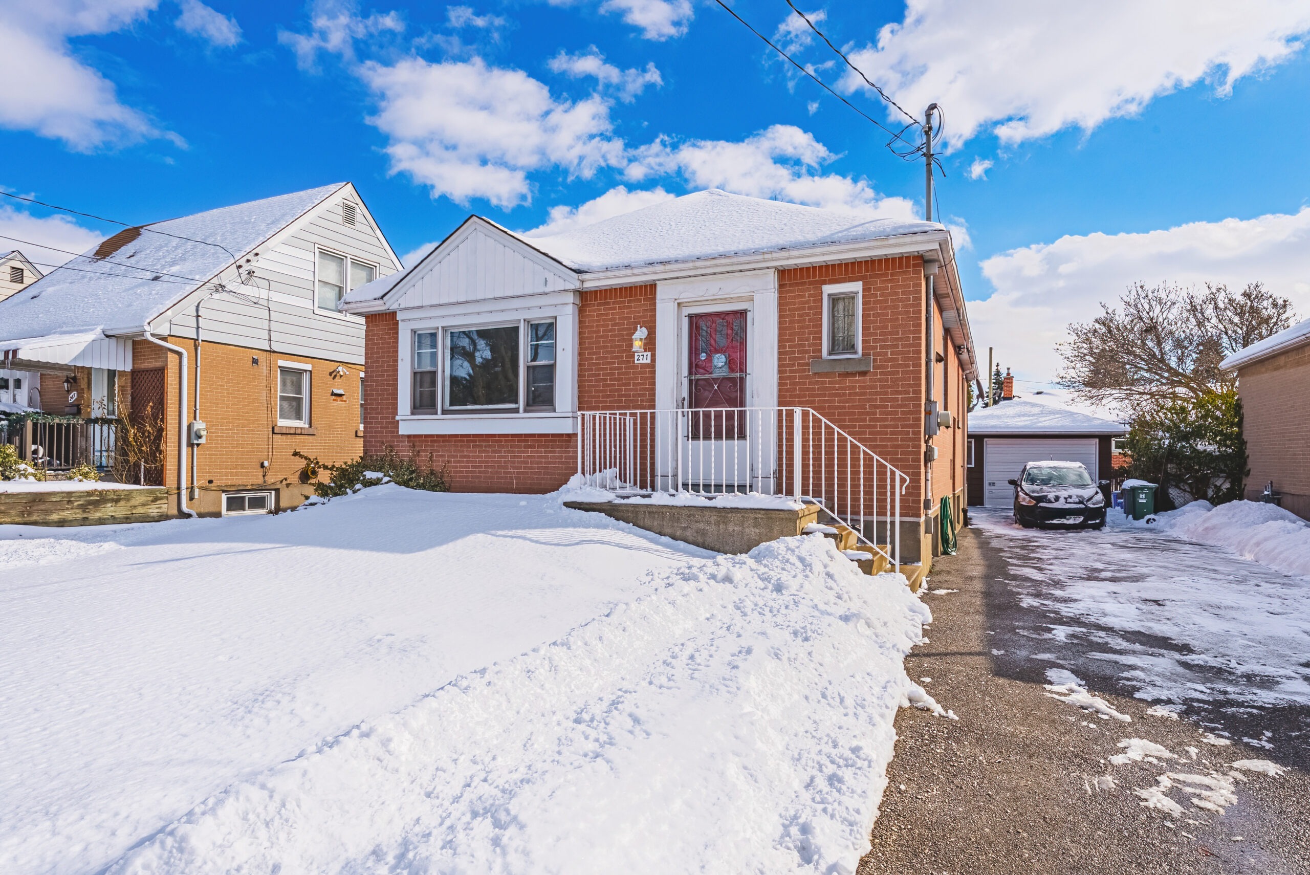 Brick house with a snow-covered front yard and stairs leading to the door, situated next to a driveway with parked cars. Clear blue sky above.