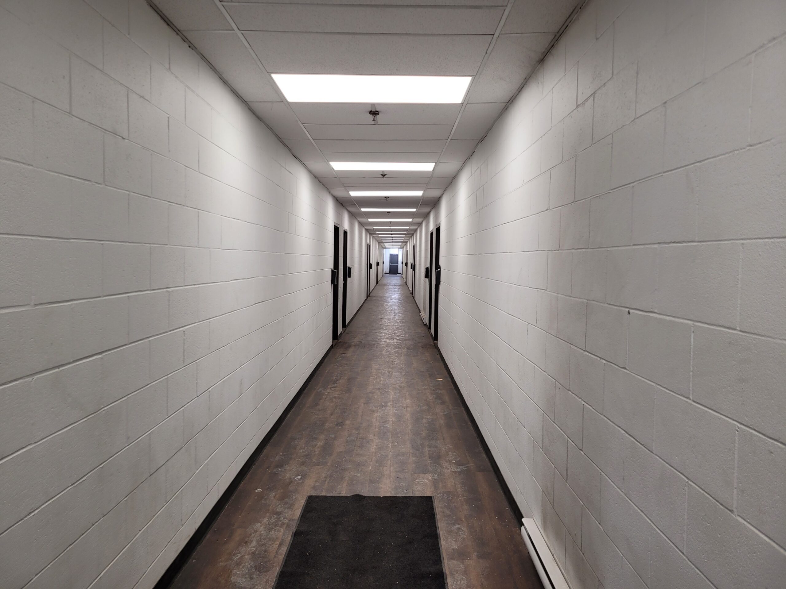 Long, empty hallway with white cinder block walls and doors along the sides. A dark mat is at the entrance, enhancing the sterile environment.