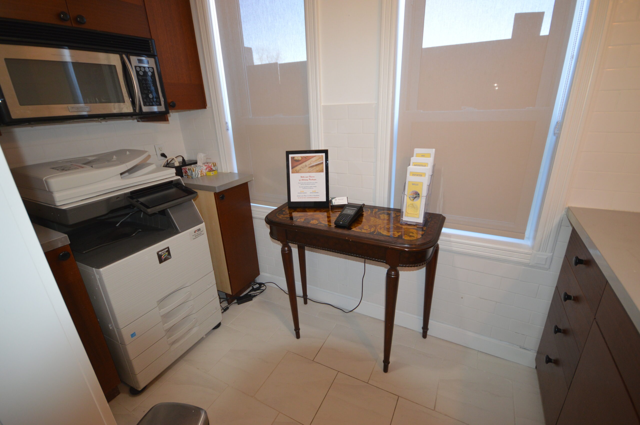 Office corner featuring a printer, vintage table with a brochure holder, and a phone. Natural light filters through the window blinds.