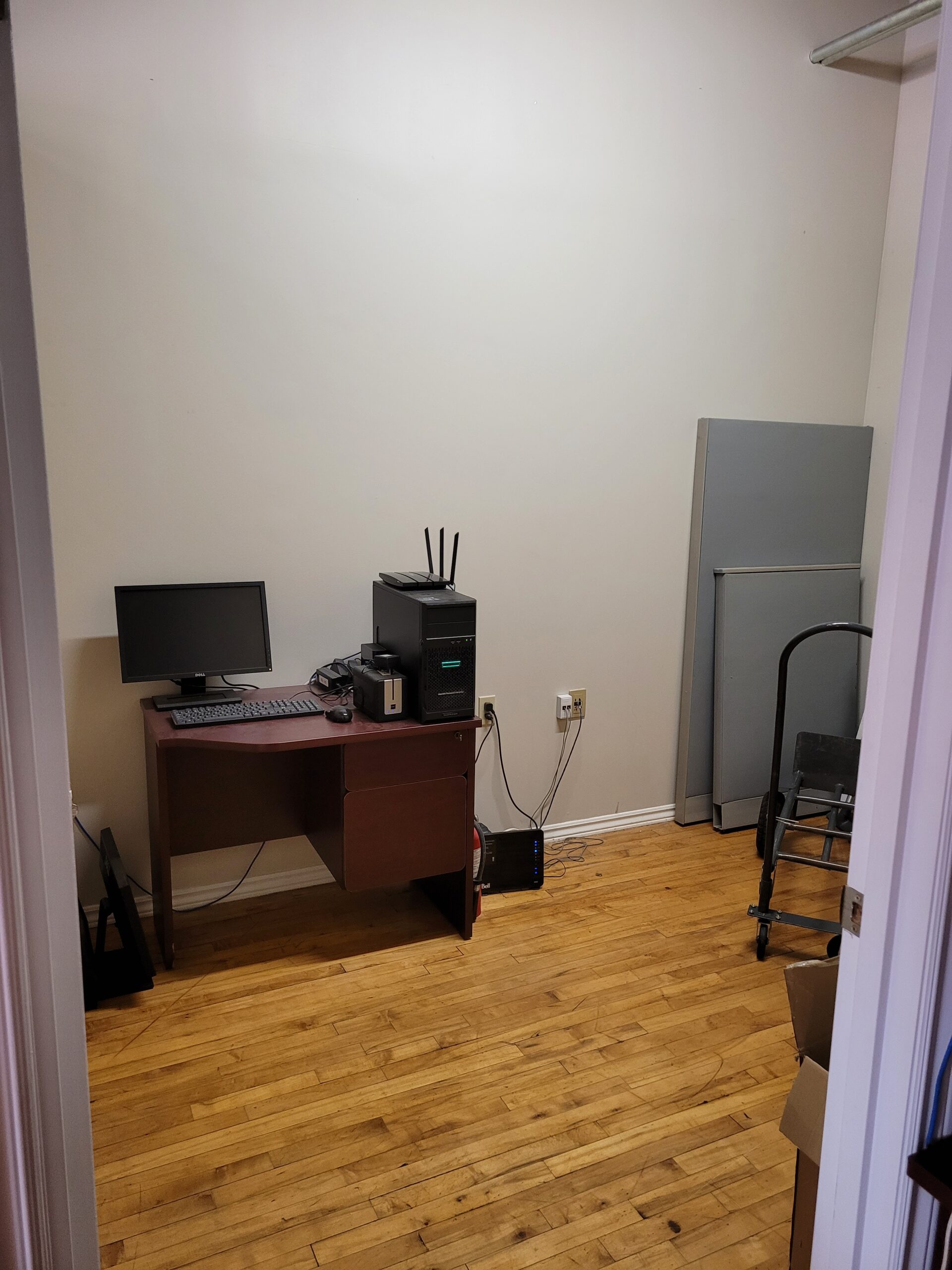 A small, sparsely furnished room featuring a desk with a monitor and computer, a router, and a nearby refrigerator. Ideal for a home office.
