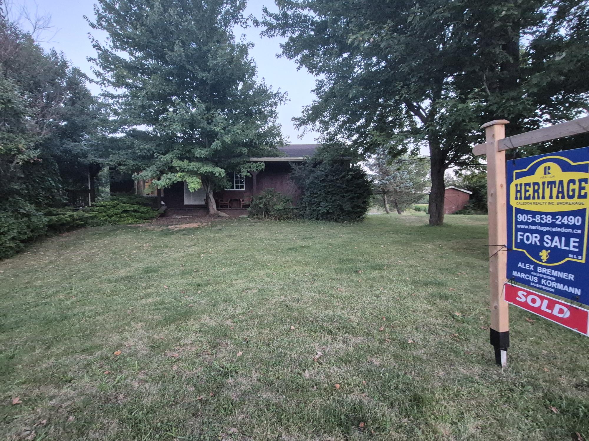 For sale sign in front of a ranch-style home, set in a grassy area with trees. The sign highlights realty details and indicates sold status.