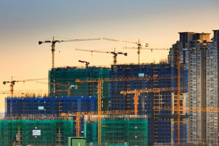 Construction site with multiple cranes and building structures, showcasing ongoing urban development against a sunset backdrop.