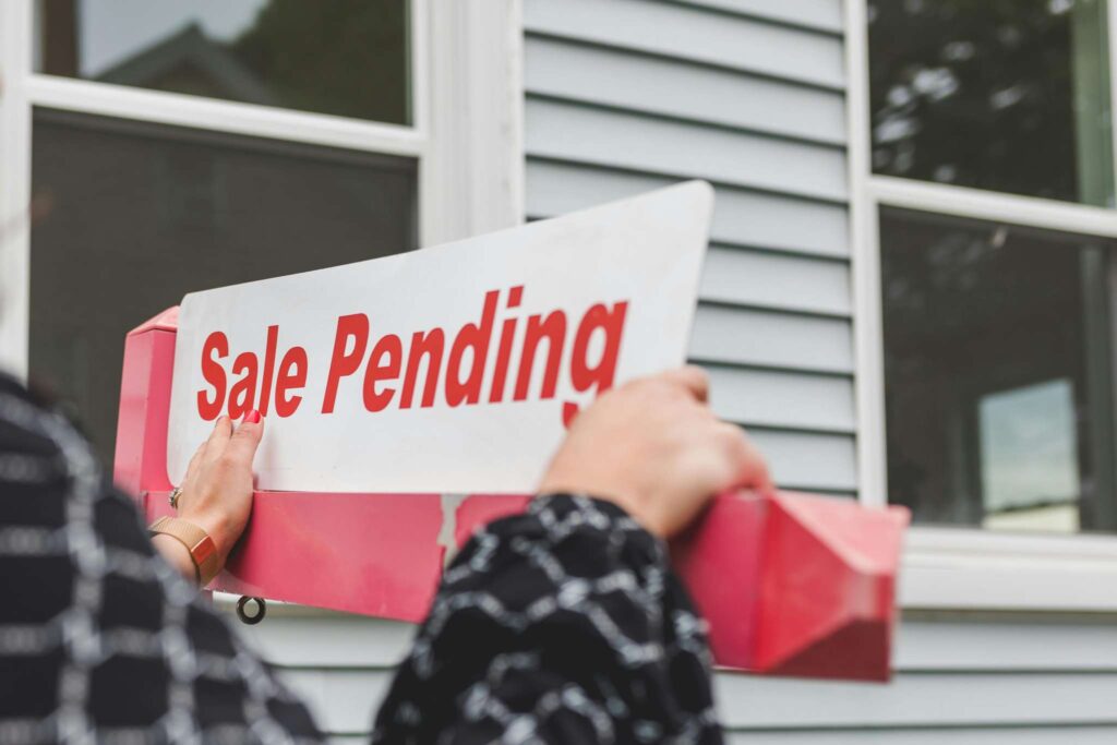 Hand holding a "Sale Pending" sign in front of a house, indicating a property is under contract. Relevant for real estate listings.