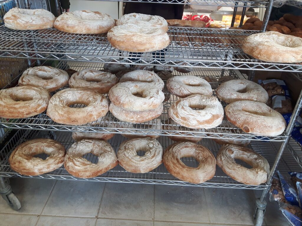 Freshly baked bagels dusted with flour, arranged in multiple tiers on wire racks in a bakery. The image highlights the variety available for customers.