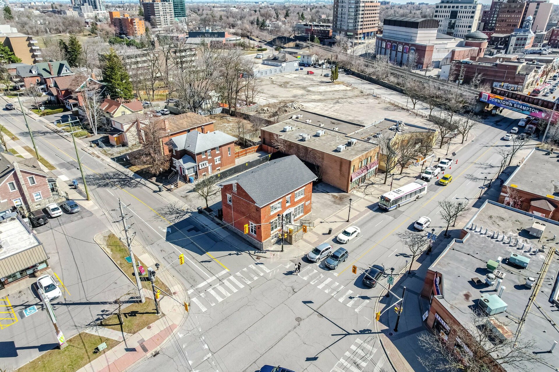 An aerial view of a downtown street intersection featuring a mix of brick commercial buildings, residential houses, parked cars, and a public transit bus on the road.