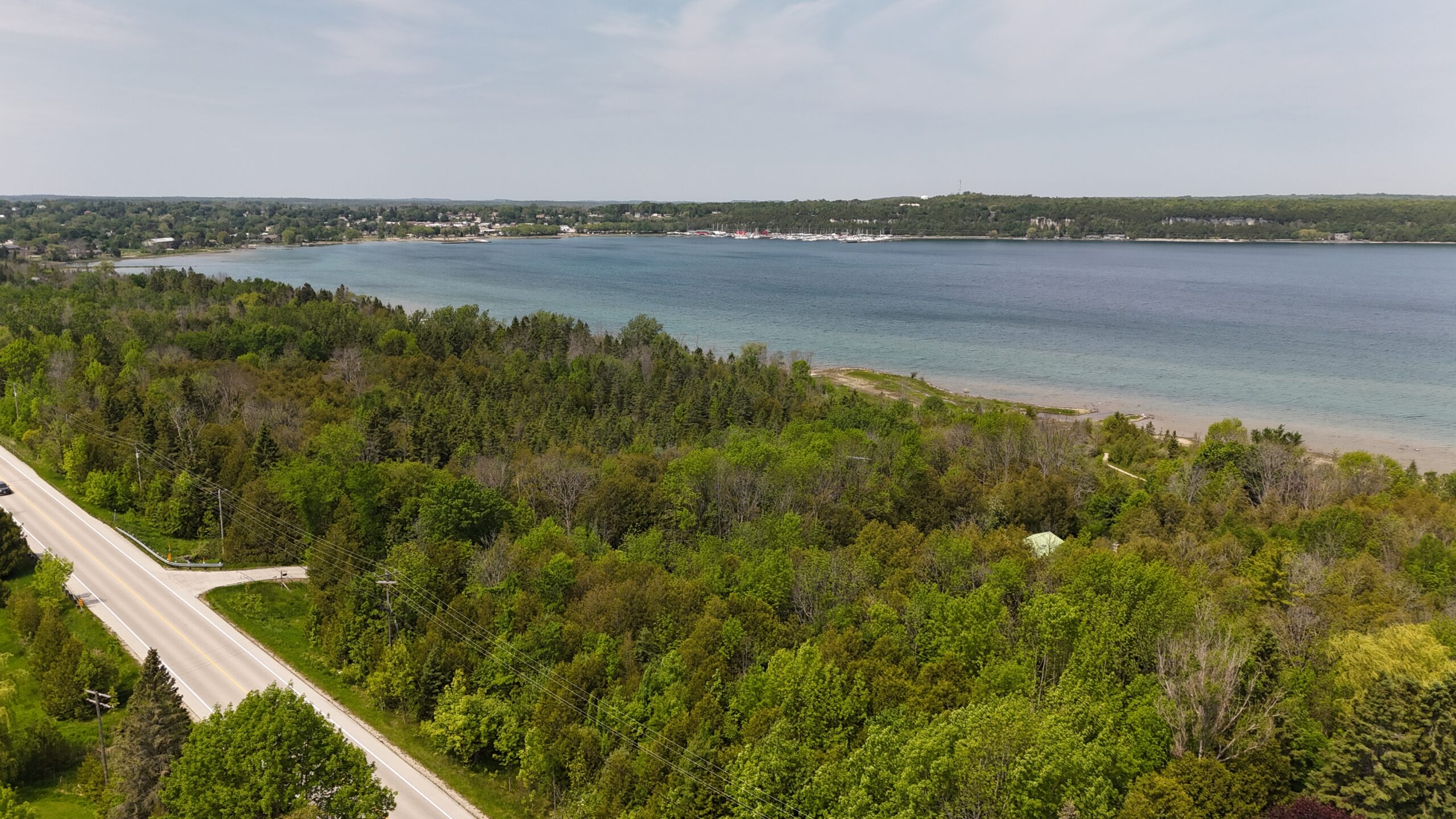 Aerial view of lush green forests along a calm lake, with a highway visible in the foreground and a small town by the water's edge.