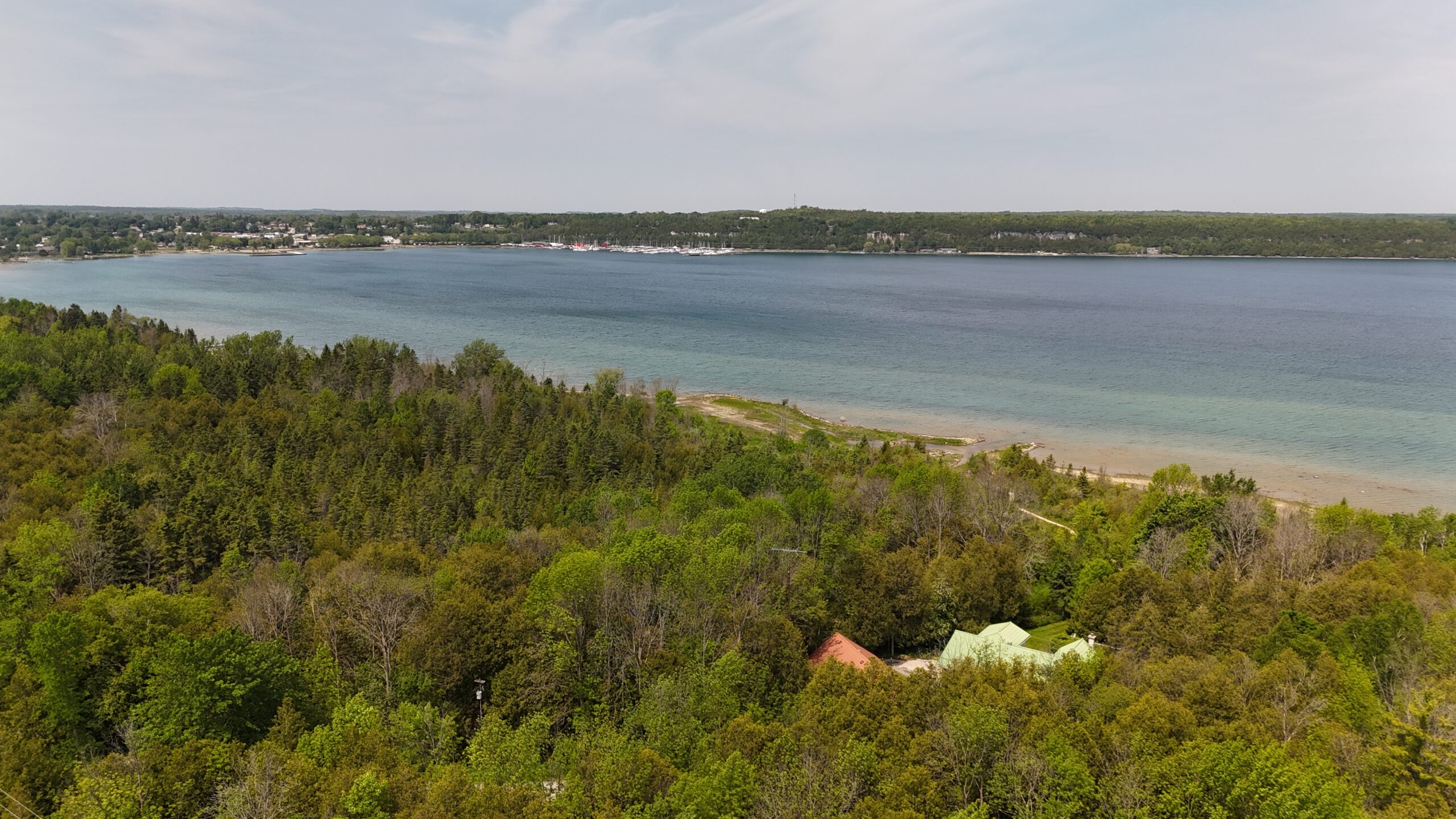 Aerial view of a coastline with lush greenery and a clear blue lake. Nearby, a small settlement and boats are visible, showcasing a serene natural landscape.