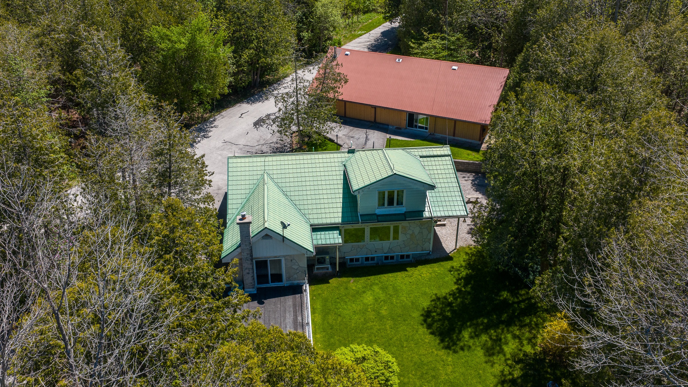 Aerial view of a modern house with a green metal roof, surrounded by trees and a gravel driveway, highlighting a spacious and serene setting.