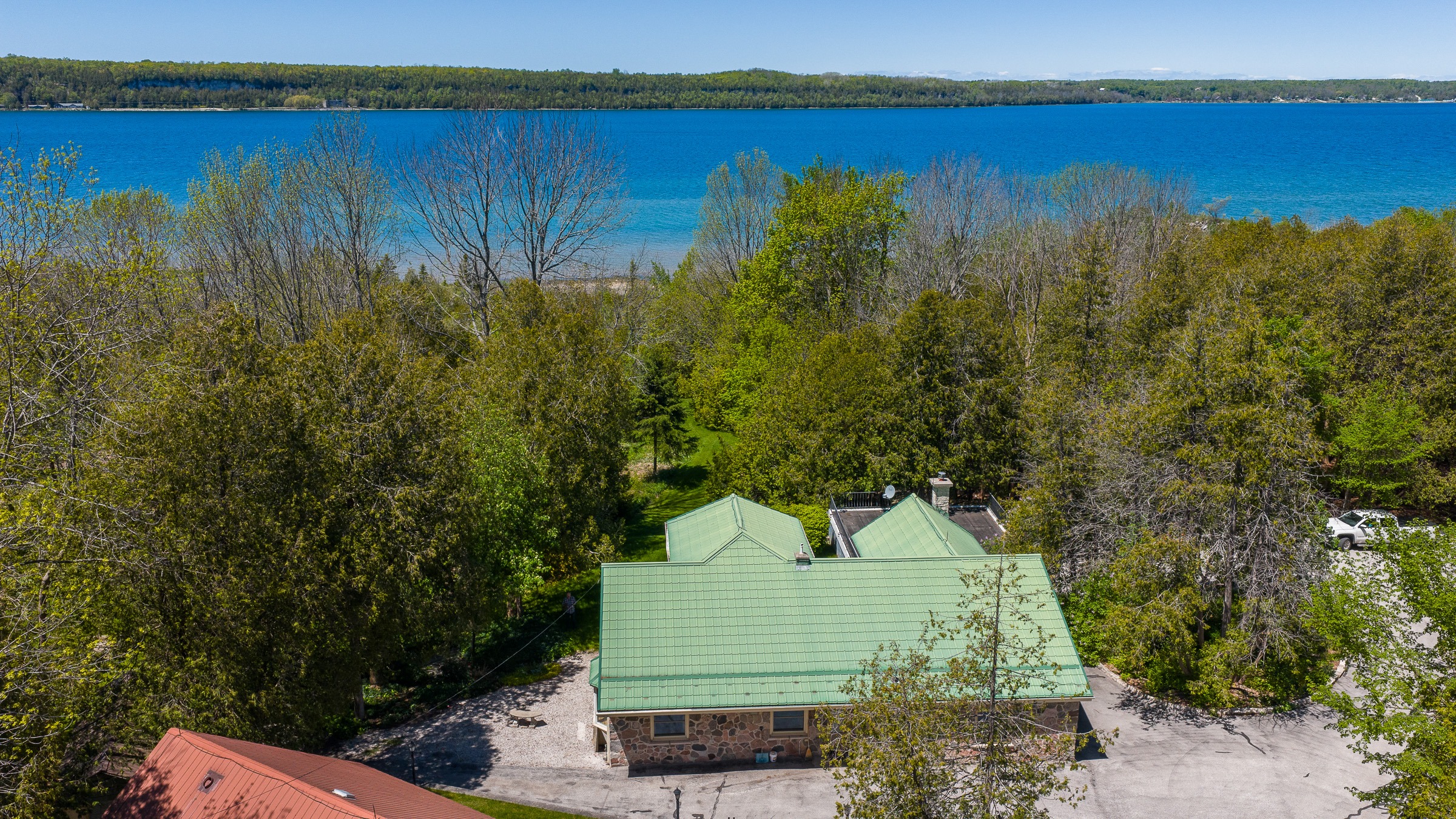 Aerial view of a green-roofed house surrounded by lush trees, overlooking a blue lake with distant hills, capturing a serene natural setting.