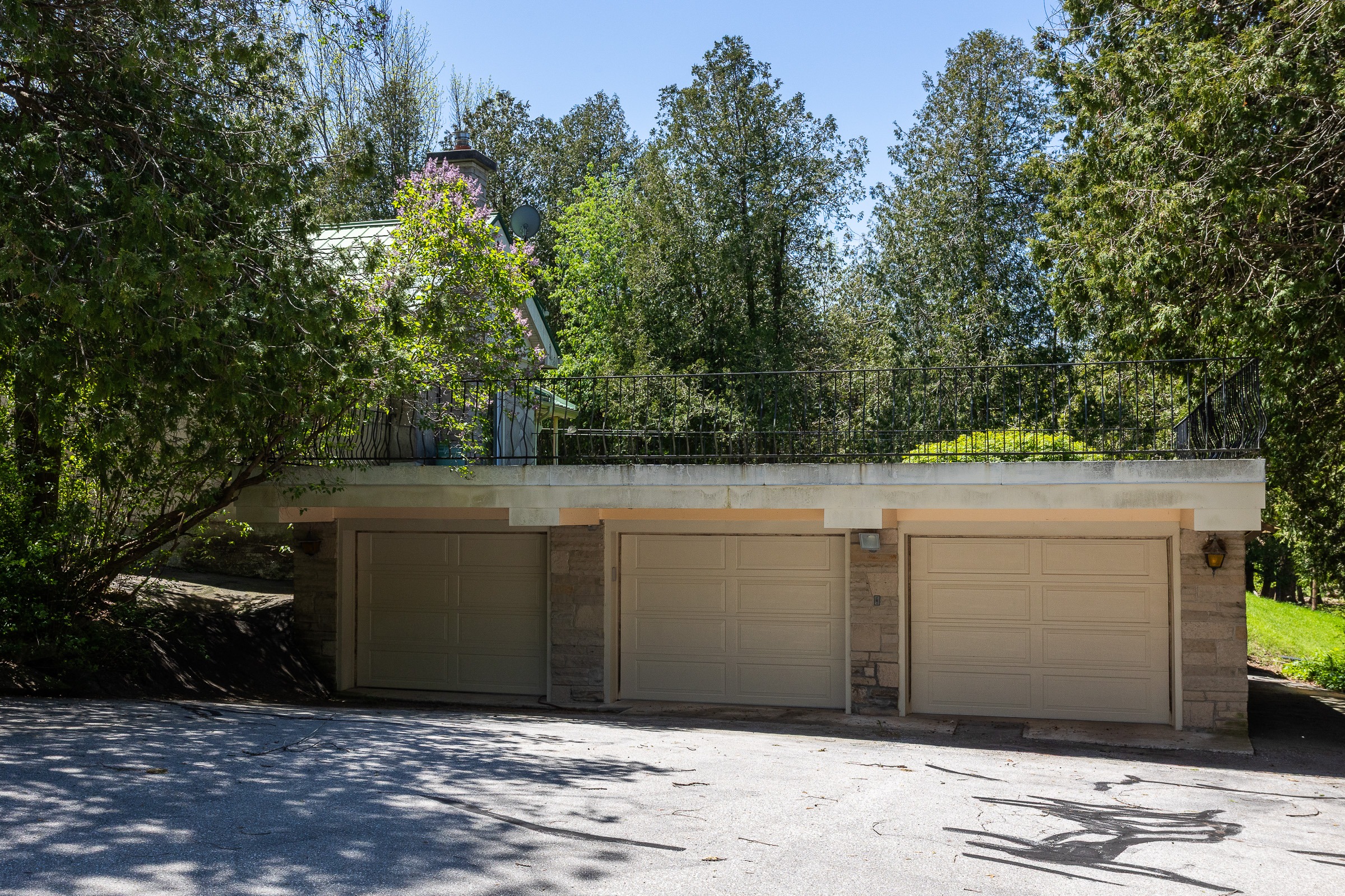 A three-garage structure situated beneath a house, surrounded by dense greenery and trees, with a clear blue sky overhead.