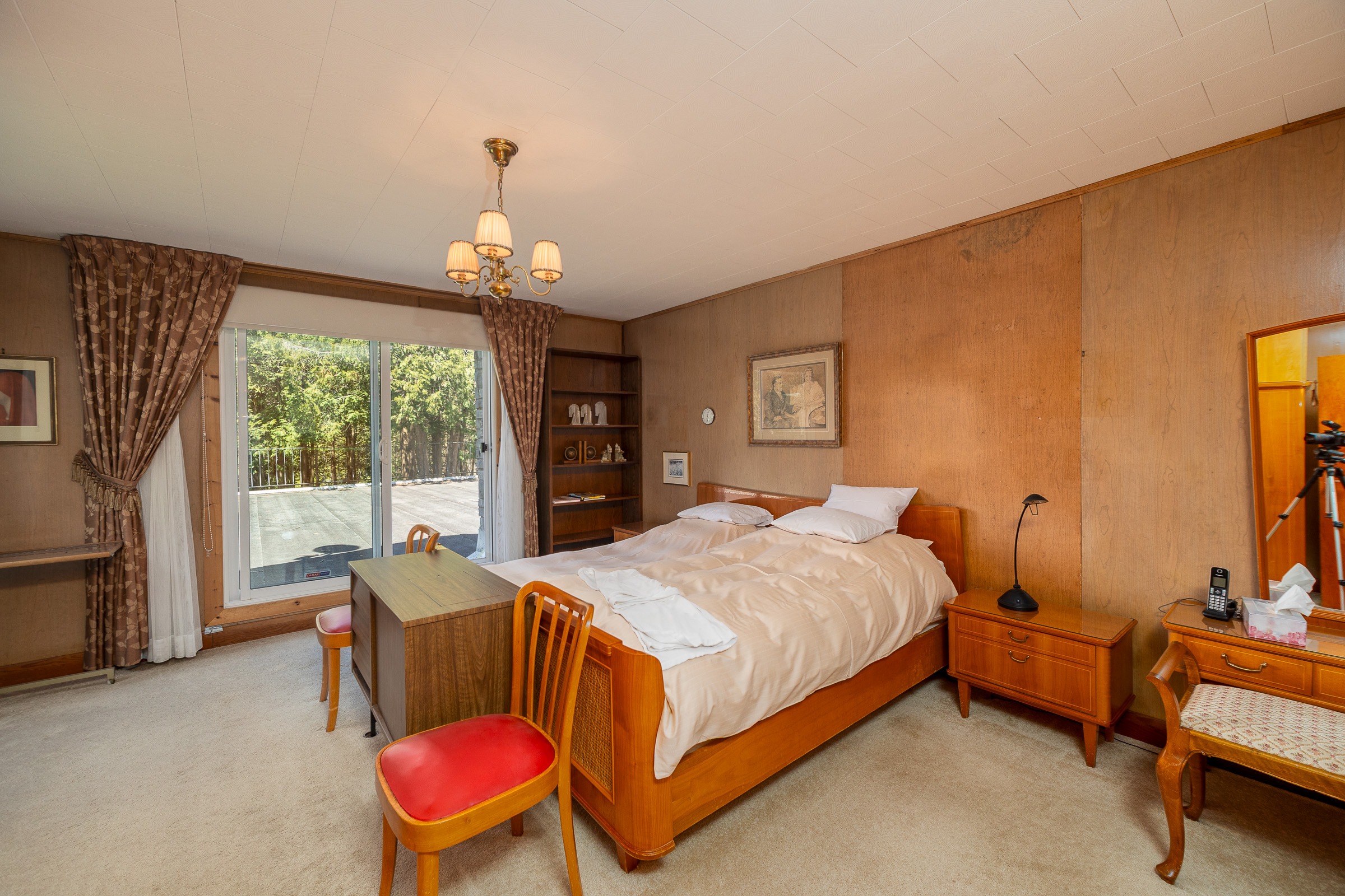 Cozy bedroom with a wooden bed, a desk with two red chairs, and large windows opening to a green outdoor view. Retro decor adds charm.