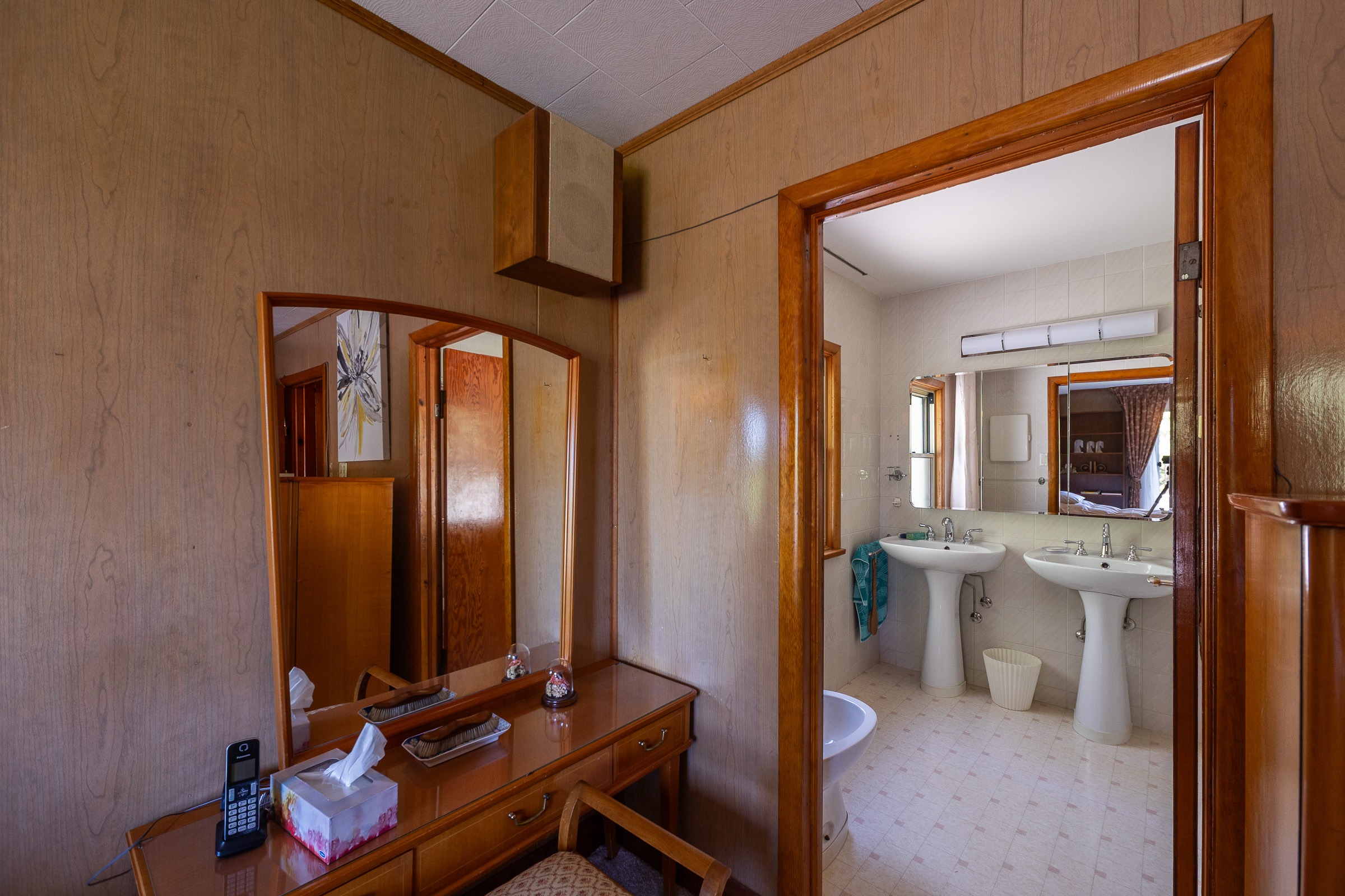 Brightly lit bathroom with dual sinks and a toilet, viewed from an adjacent room featuring a wooden vanity and patterned chair.