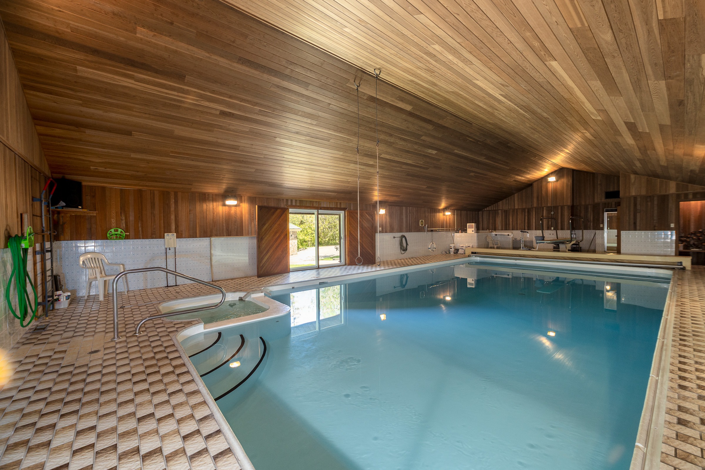 Indoor swimming pool featuring a wooden ceiling and tiled flooring, with a hot tub and nearby seating. Natural light streams through large windows, enhancing the serene atmosphere.