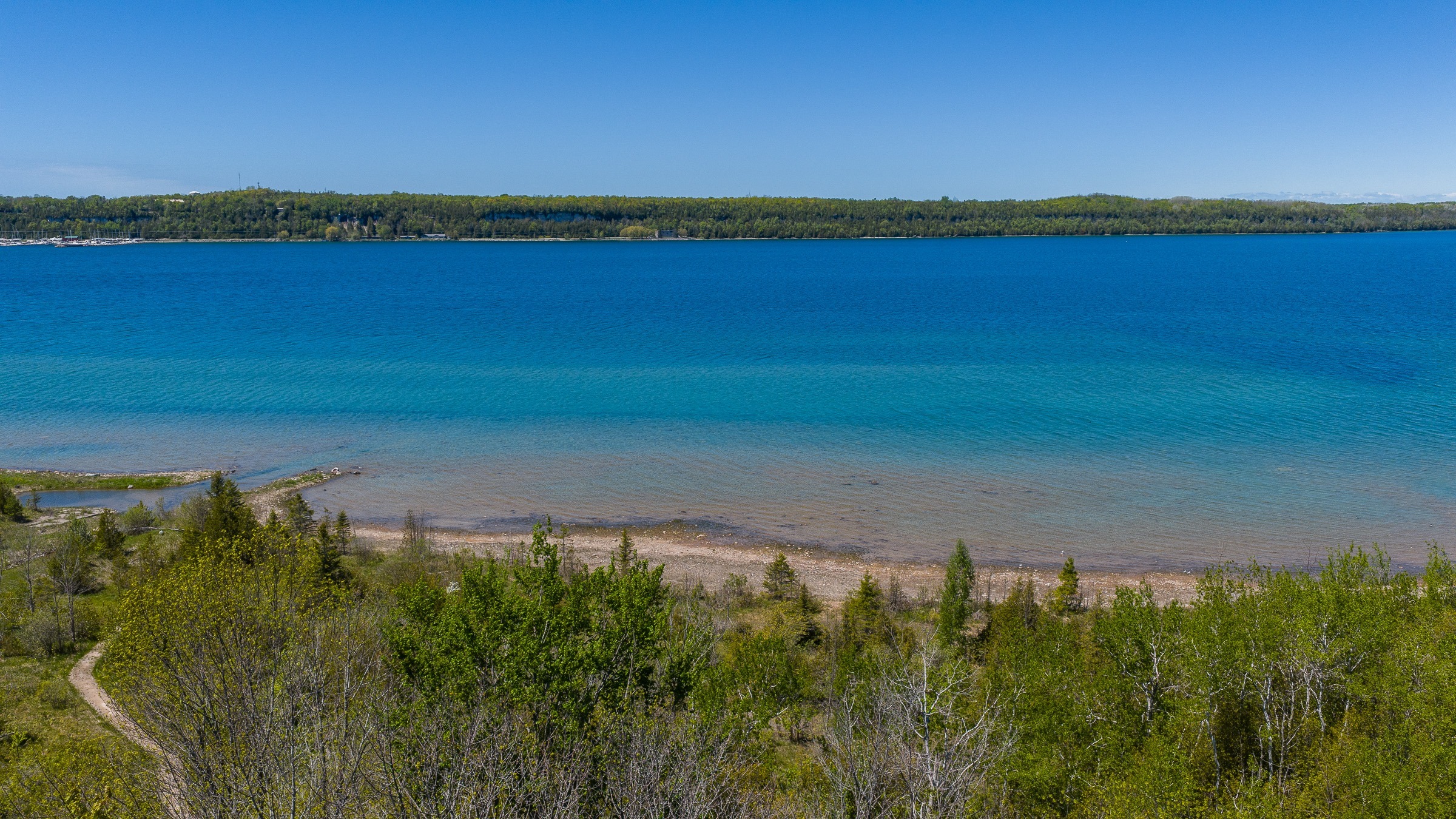 A serene view of a clear blue lake bordered by a pebbled shore and lush greenery. This landscape captures natural beauty and tranquility.