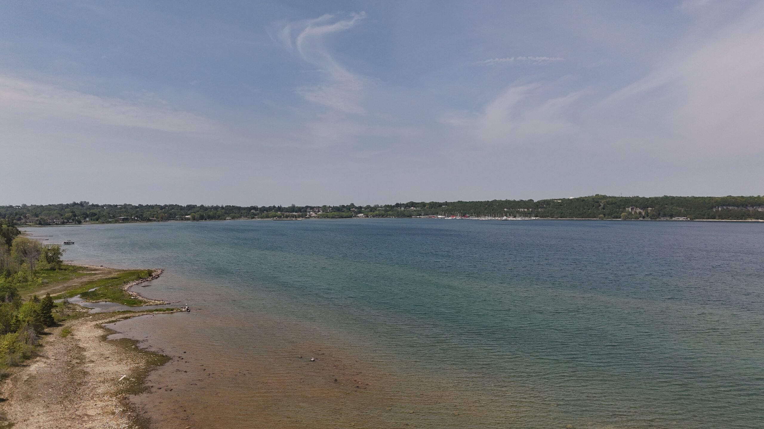 Serene waterfront view showing a calm lake with varying blue-green hues, bordered by a sandy shore and lush greenery in the background.