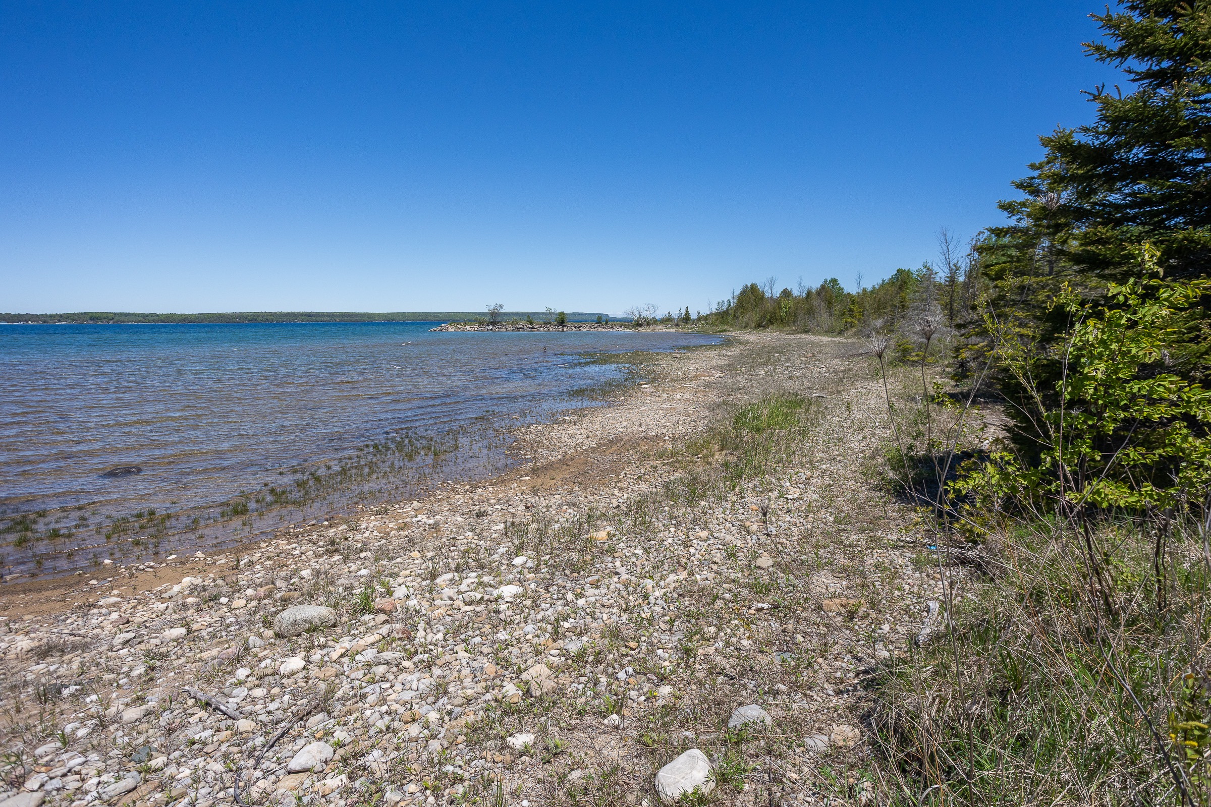 Rocky shoreline along a clear lake under a bright blue sky, showcasing the natural landscape and vegetation. Ideal for outdoor activities.