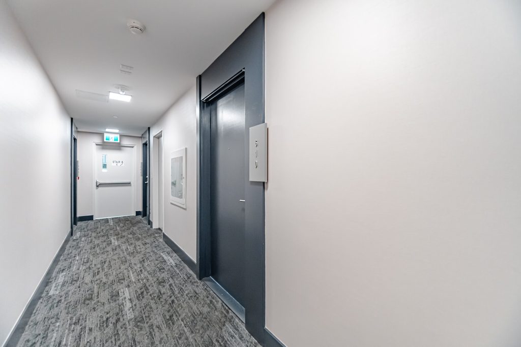Modern hotel hallway featuring a gray carpet, two elevator doors, and a wall-mounted directory, emphasizing clean and inviting design.