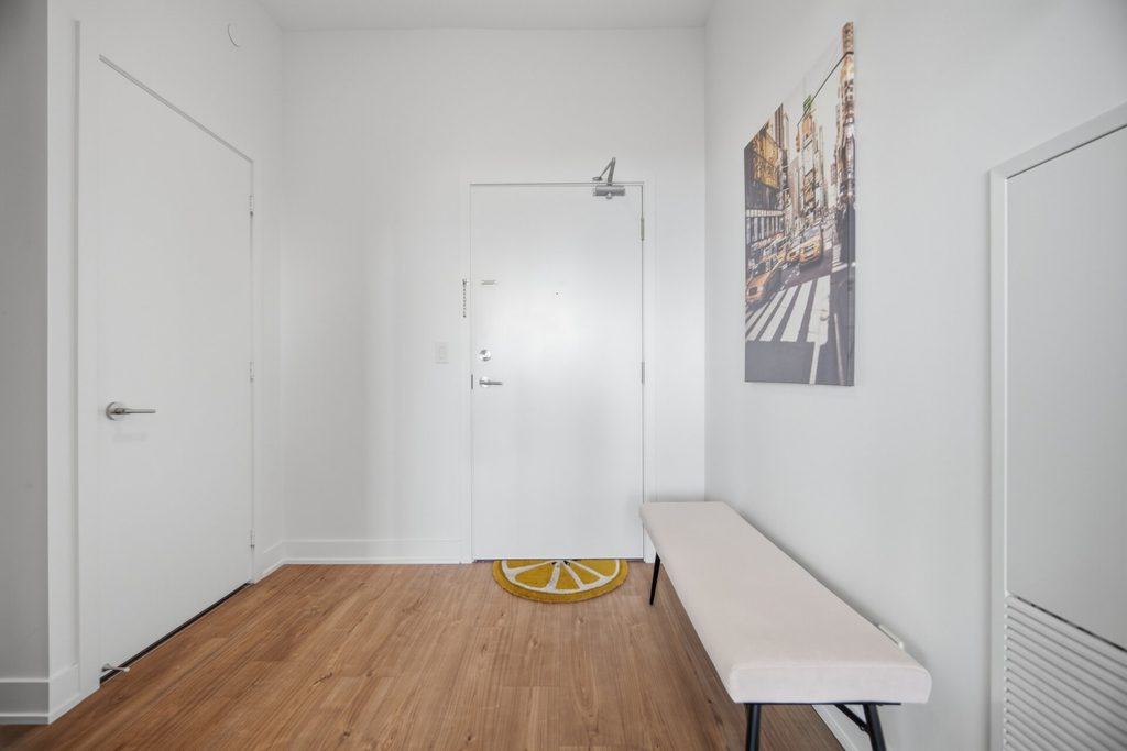 Bright, modern entryway featuring a lemon-patterned doormat, a beige bench, and a wall art piece depicting a city street scene.