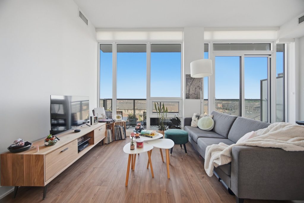 Modern living room featuring large windows with city views, a gray sofa, dining area, and warm wooden flooring. Bright and inviting atmosphere.