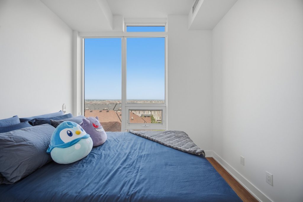Cozy bedroom featuring a blue bedspread, plush toys, and large windows offering a view of the outdoors. Bright and inviting atmosphere.
