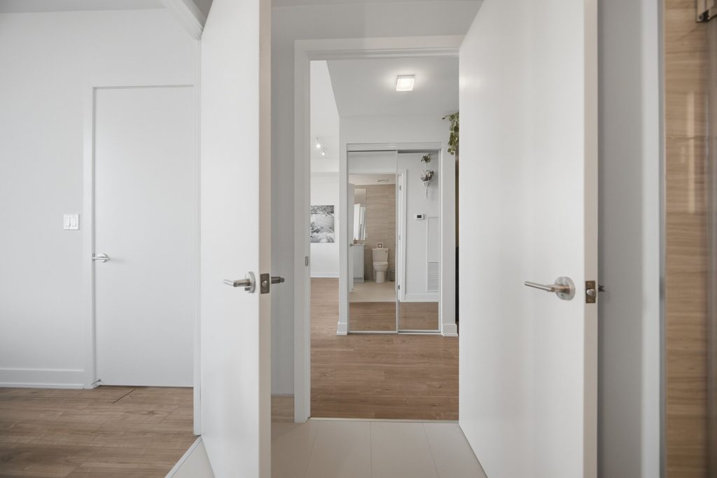 Contemporary bathroom featuring a wooden vanity with a sink, modern toilet, and a large mirror. Natural light fills the space from an adjacent room with a bed.