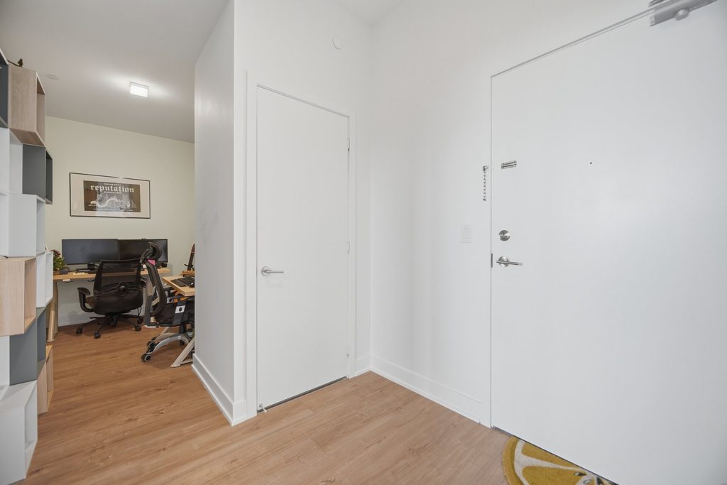View through open doors into a modern hallway, showcasing light wood flooring, a mirrored closet, and a bathroom in the background.